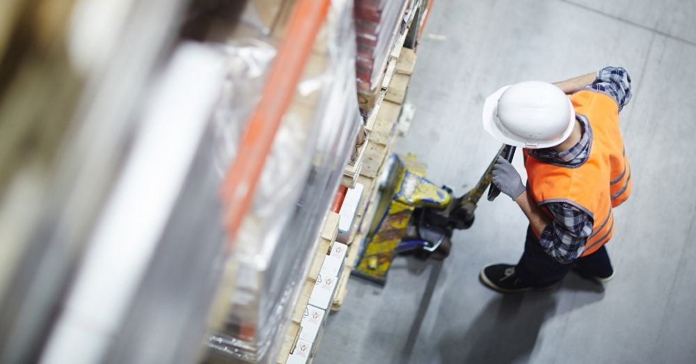 Warehouse worker in a safety vest and hard hat using a pallet jack to move goods on shelves.
