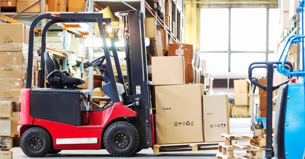 Red forklift moving stacked cardboard boxes inside a warehouse filled with inventory.