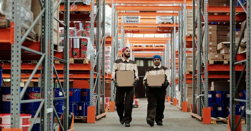 Two male warehouse workers, wearing beige and dark brown uniforms, walk down an aisle between tall shelves stacked with inventory.