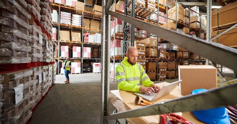 A male warehouse worker with a shaved head and a beard, wearing a bright yellow high-visibility jacket, sits at a desk and types on a laptop.