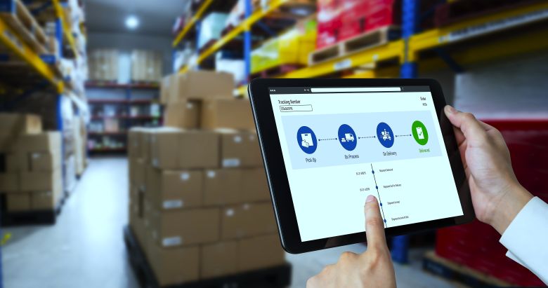Person holding a tablet showing a package tracking workflow in front of pallet racks and stacked boxes in a warehouse — warehousing services.
