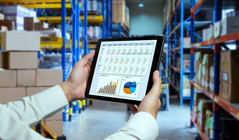 Person holding a tablet displaying inventory charts and graphs in a pallet-racked warehouse aisle — warehousing services.