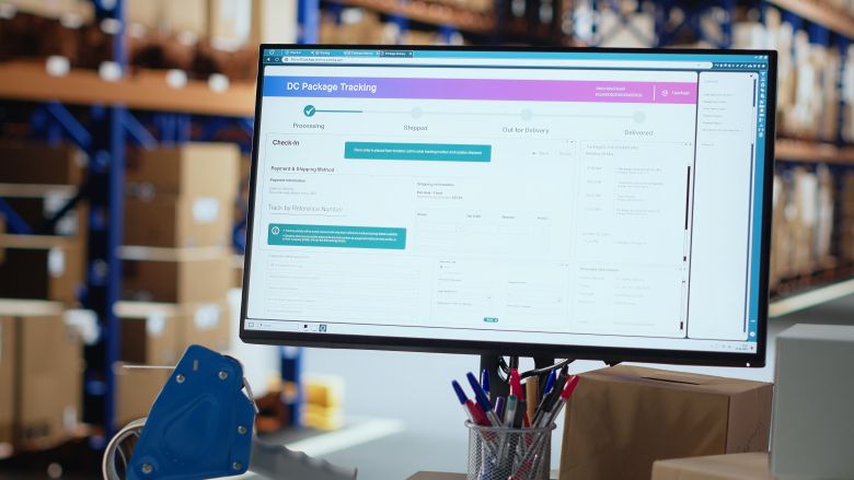 Monitor displaying a DC package-tracking dashboard on a desk in front of pallet racks and stacked boxes in a busy distribution area — warehousing services.