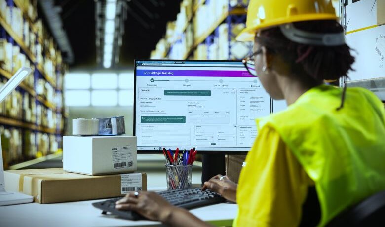 Warehouse worker in a hard hat and high-visibility vest at a desk typing on a keyboard with a DC package-tracking dashboard on the monitor and stacked boxes and pallet racks in the background — warehousing services.